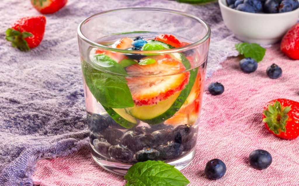 Photo showing a glass of the fruity cucumber mint hydrator, and some of the herbs and fruits that go into it.